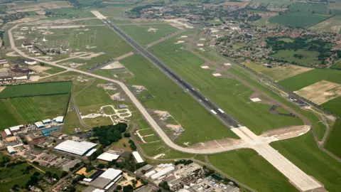 Mike Page Aerial Photogrpahy RAF Mildenhall airfield seen from the air, featuring a runway surrounded by grass and a perimeter road. An industrial estate is at the bottom of the picture, with the village of Beck Row at the top right.