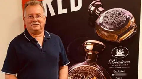 Facebook David Crisp, a man with short greying hair, a grey moustache and glasses, wearing a navy polo t-shirt and his hands clasped, stands in front of a poster showing two circular perfume bottles embossed with horses and a circular pattern and the Boadicea logo in italics. 