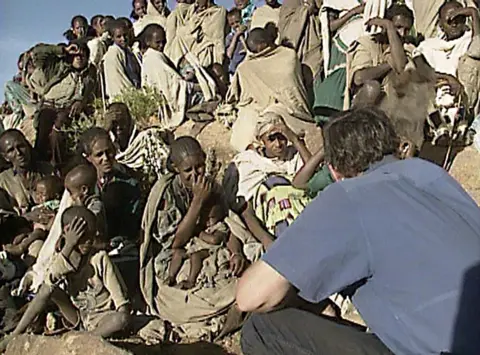 A group of Ethiopians talk to journalist Michael Buerk, who is dressed in a blue T-shirt and has his back to the camera. This is a still taken from a 1984 news report.