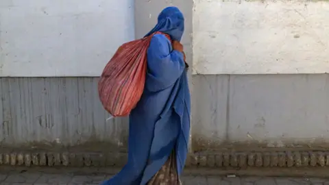 Getty Images A woman wearing a blue burqa walks down a street in Kabul with a red sack over her shoulder. You cannot see any of her face