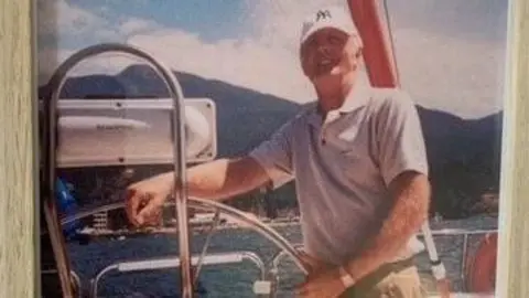 Handout An old photo of a man in a white yankees cap with mountains behind him standing on a boat on a sunny day, smiling. 