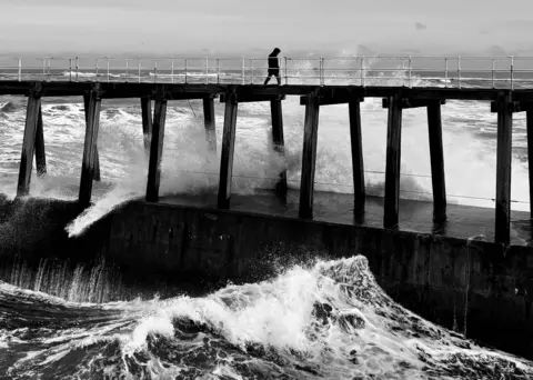 Helen C Green A figure walks along a pier over the sea as waves break below