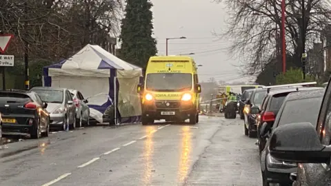 A rain-soaked road, there are park cars either side and a yellow ambulance with its lights on. It is next to a blue and white tent.