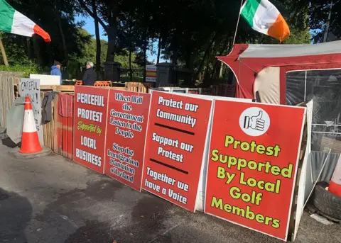 A row of four red posters with yellow and white text read: Peaceful protest supported by local business; When will the government listen to the people, Stop the immigration plantation of Ireland; Protect our community support our protest together we have a voice; Protest supported by local golf members 