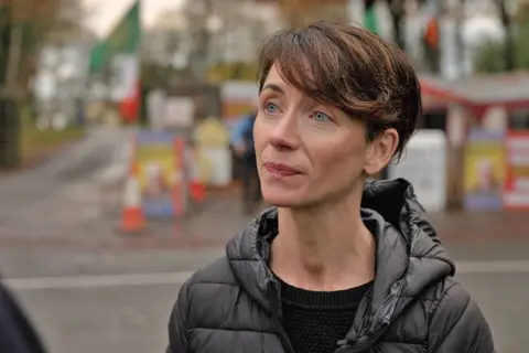 Andrea Crowe, a woman with short brown hair wears a light puffer jacket, the protest camp with signs and traffic cones in the background 