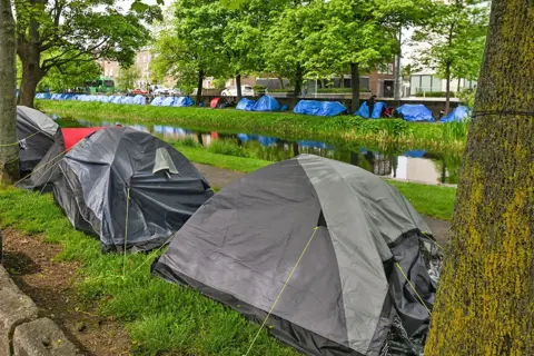 Getty Images Two rows of tents - one grey, one blue - are pitched along both sides of a canal, under trees. 