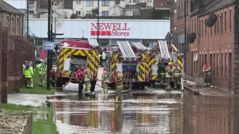 Firefighters pumping water out of Kings Row, Coalisand on Saturday.  There are three fire appliances and several firefighters in the red-brick lined street.   Some are ankle-deep in flood water. 