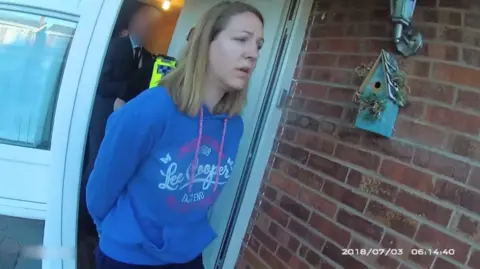 Cheshire Constabulary Police bodycam footage of Lucy Letby, who has straight blonde hair and is wearing a blue Lee Cooper branded hoodie, being led from her front door in handcuffs