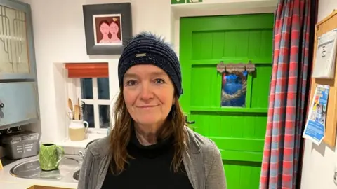 Helen Manley Jones standing smiling in the kitchen of her cottage. She is wearing a dark blue hat and has long brown hair and wearing a grey casual jacket over a black top. Behind her is a light green door and a small window. To her left is a silver metal sink with a green patterned ceramic jug on the drainer.