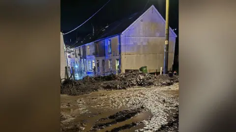 Jack Humphrey A picture showing mud strewn across a street. The photo is taken from the top of the street, in the foreground there is loads of mud and sludge, and you can see houses towards the back of the picture on a street where there is the flashing blue of the emergency services. 