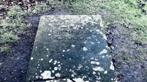 A gravestone, set amid a muddy patch of ground, reads Ebenezer Scrooge. There is lichen growing on its surface and grass at the top of the image.