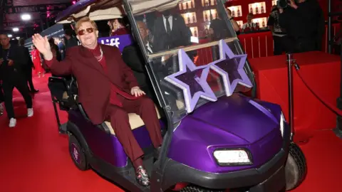 Getty Images Sir Elton waving and riding in a golf cart-type buggy