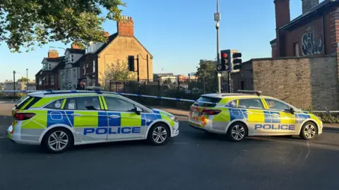 Alice Cunningham/BBC Two police cars in front of a police cordon blocking a road in Ipswich. A row of terraced houses can be seen in the background. 