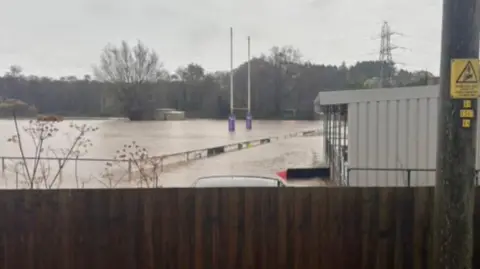 Newport High School Old Boys RFC A rugby pitch under a few feet of dirty water. You can see rugby posts in the distance and the top of metal railings around the pitch. There's a brown fence in the foreground there are trees beyond the pitch.