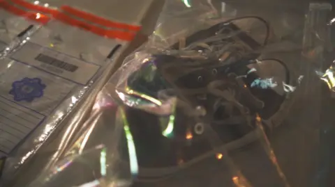 Plastic bags on a table contain some of Kelly's belongings after being returned to her parents. A large bag on the right contains a pair of black low top trainers with a white rubber sole and white rubber tip at the toe. There are white laces and small metal hoops visible, where the shoe would be tied. A smaller bag, with orange tape at the top, bears the Garda emblem and a barcode. It's placed to the left of the shoes and contains a small collection of piercing jewellery. 