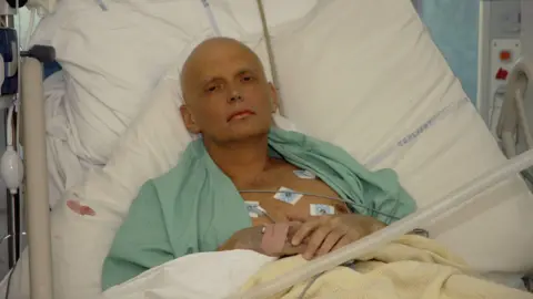 Getty Images Alexander Litvinenko lying in a hospital bed on an intensive care unit. Lots of wires are linked up to his chest. He has a bandage on one hand. He is looking into the camera with a blank expression on his face. 