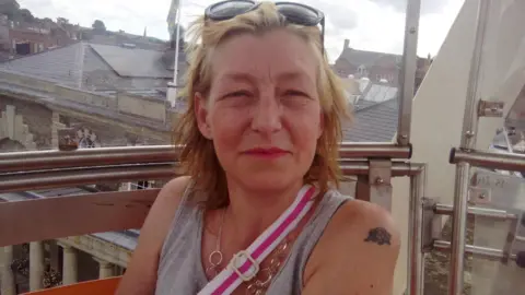 Facebook Dawn Sturgess looking at the camera. She has blonde hair and is wearing a grey vest top. She has sunglasses on her head and the strap of a bag over her shoulder is visible and is wearing a necklace.