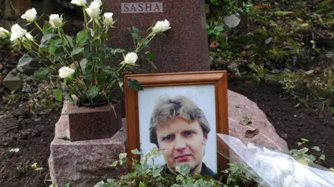 PA Media Alexander Litvinenko's grave. A framed photograph of him is in front of the headstone and there is a vase of white roses. The headstone has a plaque reading "Sasha" on it - a nickname for him.