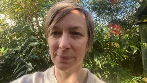 Fiona Button Selfie of woman in a garden, head and shoulders only. She is 48, has blonde hair and is wearing a beige wool jumper.