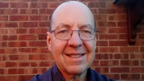 Andrew Bullock A selfie of a smiling man standing in front of a brick wall, outside. He is 60, bald and wearing glasses.