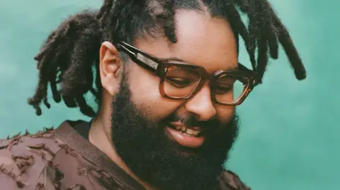 Toby Coulson Elmiene pictured looking down and smiling. He wears a brown top and square tortoiseshell-rimmed glassed. He has a thick beard and wears his hair tied back and stands in front of a blue backdrop. 