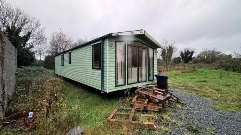 A green mobile home in a rural area with weeds growing nearby and wooden pallets beside it.