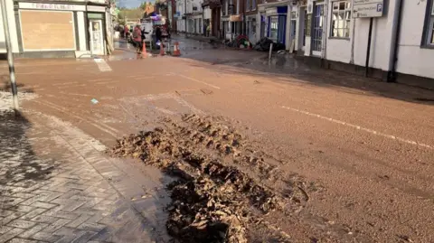 A damp street with layers of mud and rutted tire tracks. Cones can be seen blocking the entrance to a road ahead, a boarded up window by shop sign Pitter Patter, and residents talking to a reporter with a microphone. A fire engine is also visible in the background. debris is piled outside buildings lining the right hand side of the street. The Town in the Orchard pub is in the foreground on the right hand side.