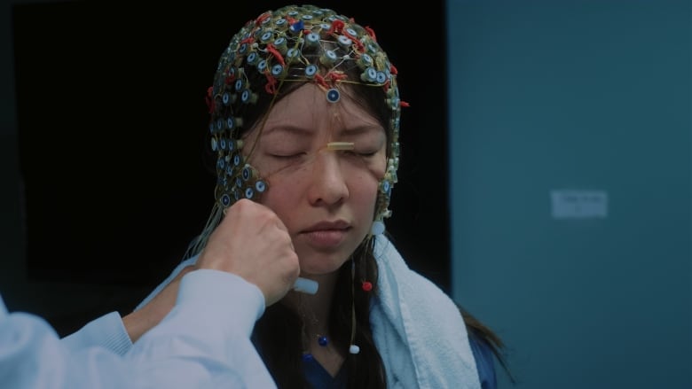 The image shows a person wearing a multi-electrode cap, often used for EEG (electroencephalography) or other types of brain activity monitoring. The electrodes are attached to the scalp using wires. A hand is visible adjusting something on the setup near the face.