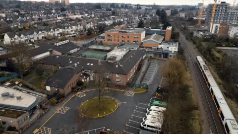 An aerial view of Whitefield School in north-east London, showing a two-storey red-brick building with a more modern extension at the rear. A London Overground line runs next to the site, with new tower blocks beyond it, while on the other side are streets of semi-detached homes.