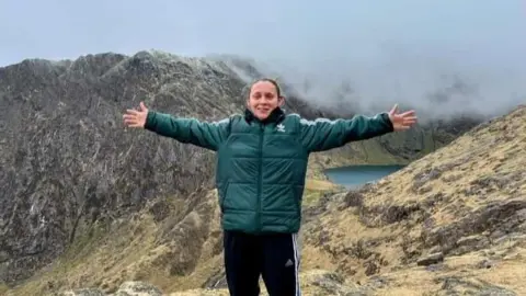 Elissa Hubbard in a green sports jacket and black Adidas tracksuit bottoms stands at the top of a mountain and a lake is also visible in the background. She has her arms outstretched and is smiling 