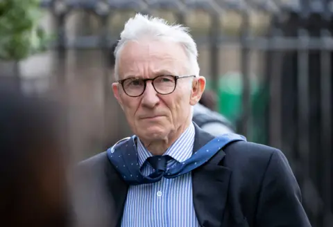 PA Media Lord Brodie walking on a street with railings behind him. He is wearing a dark blue suit, striped shirt and glasses. The wind has blown his tie over his shoulders.