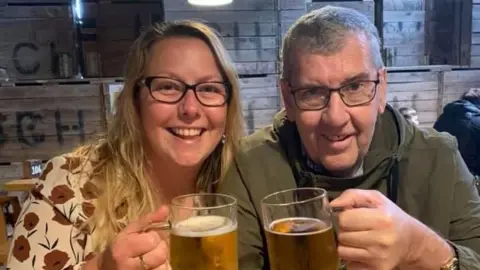 Maureen (left) and Tony Dynes in a pub, smiling at the camera and holding up pints of beer. Maureen is wearing a patterned cream top with red flowers and Tony is wearing a green jacket. Both are wearing glasses.