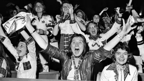 Getty Images A crowd of Roller fans scream in delight as their idols perform on stage, all wearing bits of tartan with their hands in the air. The girl at the front is wide-eyed and open-mouthed in excitement as she raises her hands in the air.