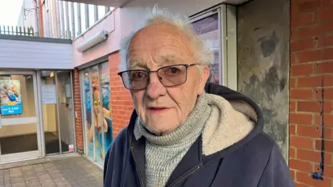 Kate Bradbrook/BBC A man with glasses, and roll-neck grey jumper. He is also wearing a hoodie. He is stood in front of a leisure centre.