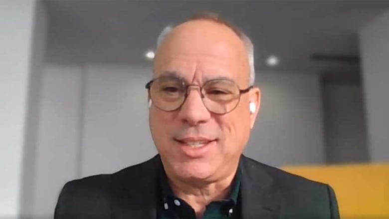 A portrait of a man with greying hair, wearing a black collared shirt, black blazer and glasses.