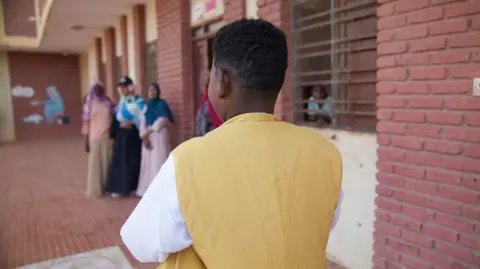 Joyce Liu / BBC The back of Mahmoud, a teenager in a yellow and white top, standing next to a building with three women stood in the distance, just out of focus