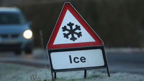 Getty Images A stock image of a ice warning road sign - a white triangle with a red border showing a black snowflake icon in the middle with Ice written below. The sign is placed on the side of a frosty grass patch. In the background is a blurred silver car driving towards it.