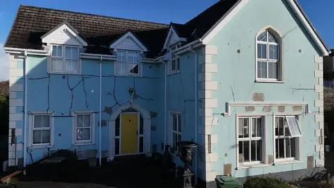 A landscape still image of a house which has been build using the defective Mica concrete bricks. A sky blue, two-storey house has a number of dark cracks across its surface. There is a yellow door on the left of the house and is surrounded by white windows.