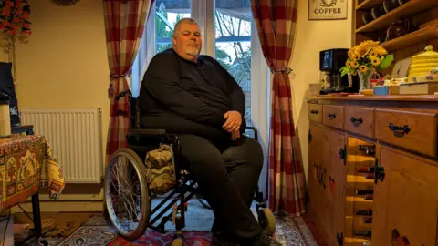 Neal Wylde Neal Wylde, wearing all black, is sat in front of French windows in a kitchen