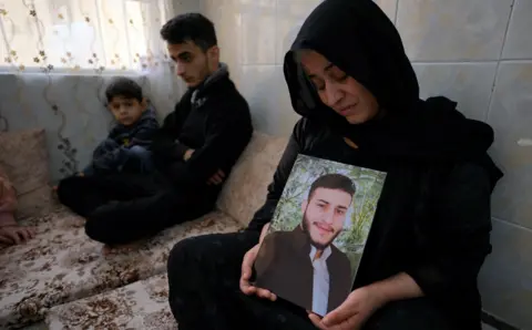 Lee Durant/BBC Shukria Badar looking sad sitting on a sofa with a young boy and young man while holding up a portrait of her son Baryar Mohamed, who died while crossing the Channel.