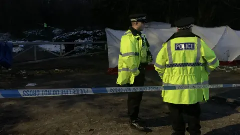 BBC/Phil Cunliffe Two police officers in hi-vis jackets stand behind blue and white police tape which reads 'police line do not cross'. Behind them is a white forensics tent on some scrub land with trees in the background.