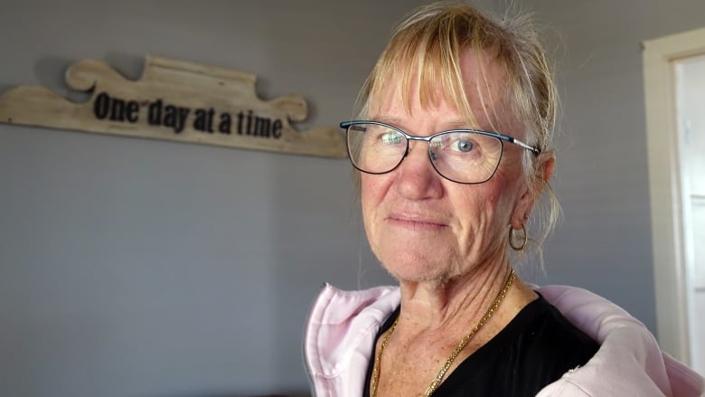 A woman wearing glasses and a pink hoodie stands in a grey room. Behind her is a sign on the wall with the words "One day at a time."