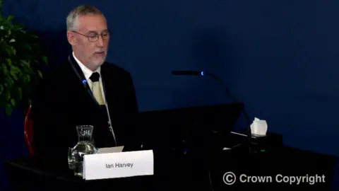 Thirlwall Inquiry Ian Harvey, with short grey hair and light grey stubble, sits at a desk containing microphones and a computer screen