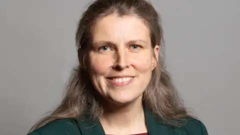 UK Parliament A woman with long light brown hair wearing a dark blazer. She stands in front of a grey background.