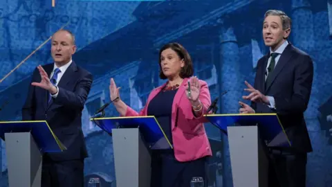 PA Media Micheál Martin stands to the left in front of a grey podium. He is wearing a blue suit and tie and is holding his hand out. Mary Lou McDonald stands in the middle in a blue top and bright pink blazer, holding two hands up. Simon Harris stands behind a podium to the right and also appears to be in the midst of talking. 