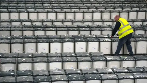 PA Media A man in a yellow hi-vis jacket is walking among a sea of grey ballot boxes, checking the lids are secure.