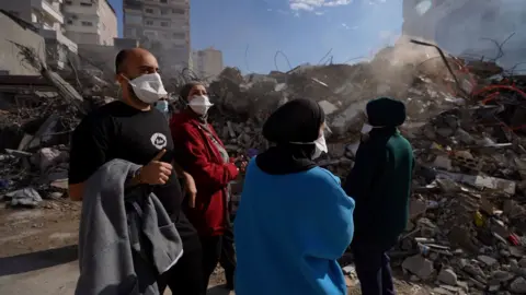 Man (L) and three women in hijabs, look at the piles of rubble around them. All four are wearing white surgical face masks.