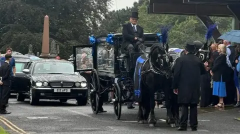 A horse-drawn carriage brings Jay Slater's coffin at the head of a cortege of cars to the chapel, with attendees lined up 