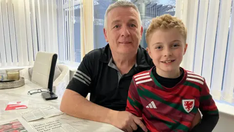 BBC News Angharad James' father sits with a child in a Wales football shirt at a table with cuttings which mention Angharad and the Wales team