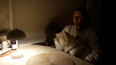 BBC/Joe Phua Nataliya sits in a chair in a poorly lit room, with a white dog on her lap